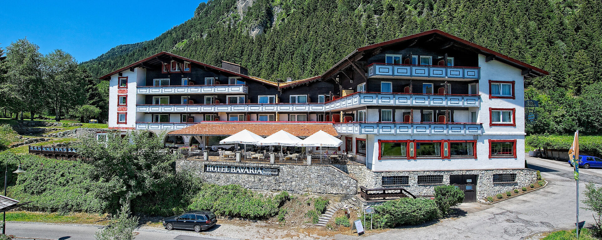 Das weiß-rote Gebäude des Hotels Bavaria Pfronten in einer Alpenlandschaft.